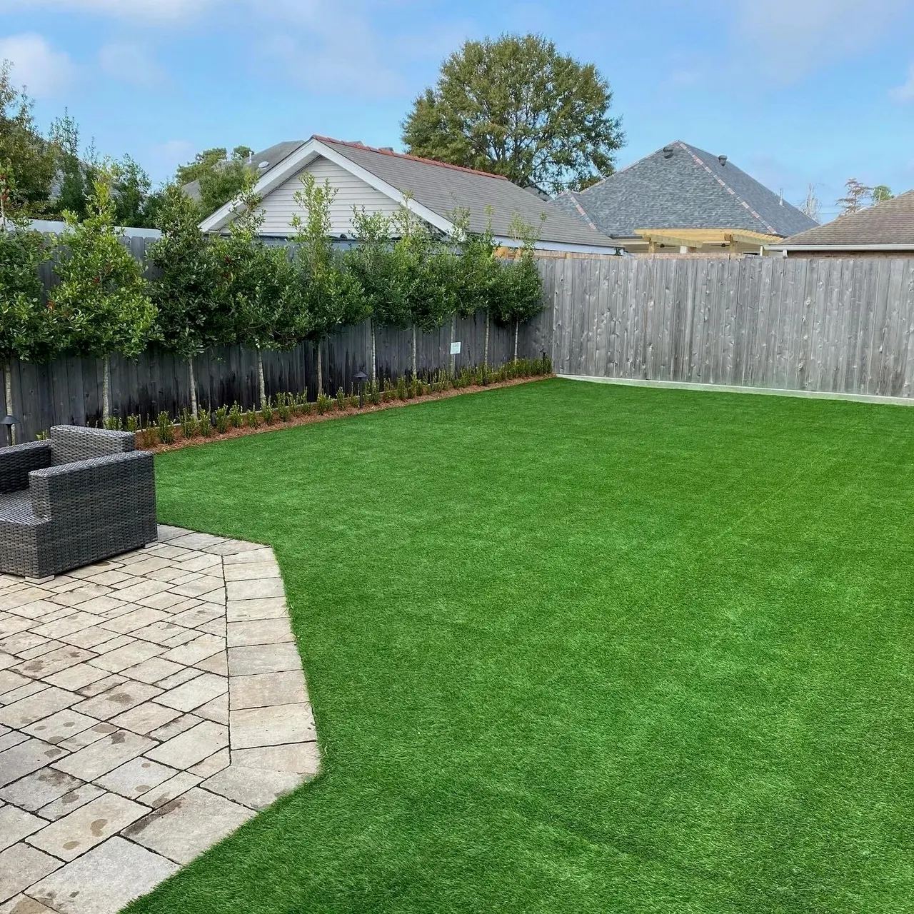 Artificial turf backyard bordered by a curved stone patio, shrubs, and wooden fencing in Fountain Valley